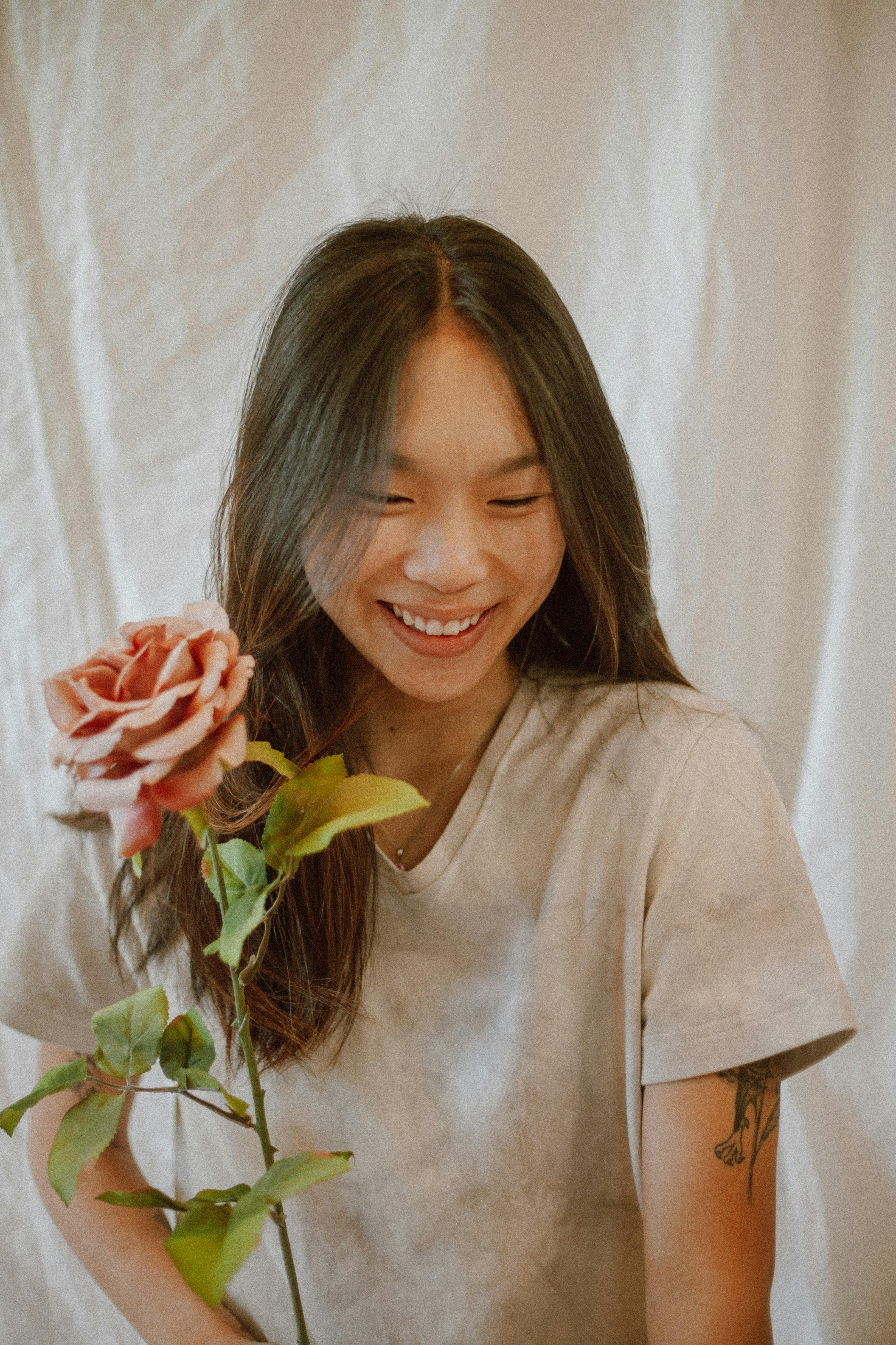 Delighted ethnic female in casual outfit with pink gentle flower with green stem and leaves smiling