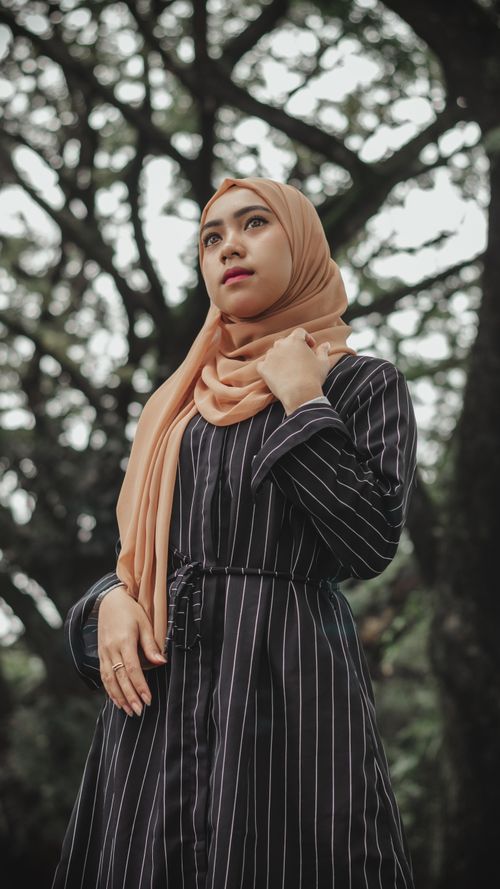 Free From below of charming Muslim female in black dress and headscarf standing in park and looking away against trees in daytime Stock Photo