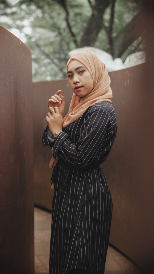 Free Side view pretty Muslim female in maxi dress and traditional headdress holding hands in front and looking at camera while standing on narrow outdoor passage Stock Photo
