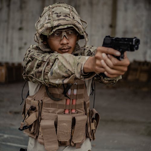 Concentrated ethnic fighter wearing camouflage uniform aiming gun against grunge concrete wall in army shooting range