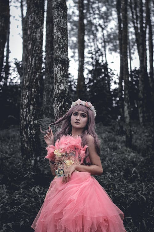 Unemotional young female wearing elegant pink gown and wreath touching hair and looking away thoughtfully while standing in green woodland