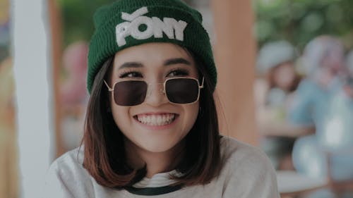 Crop delighted young female in hat and trendy sunglasses standing on street and looking away with toothy smile