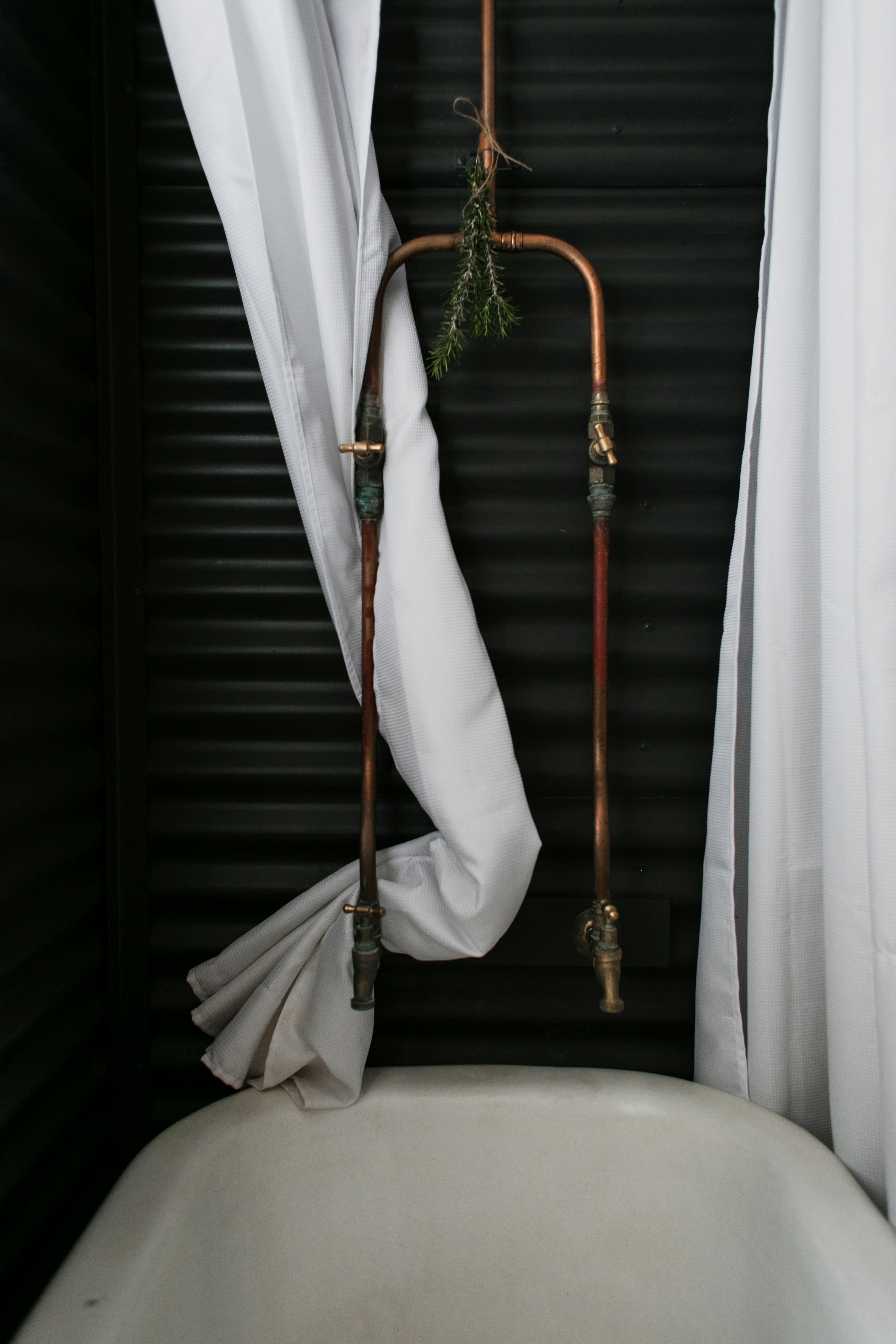 Rustic bathroom features exposed copper pipes and white curtains for a chic minimalist look.