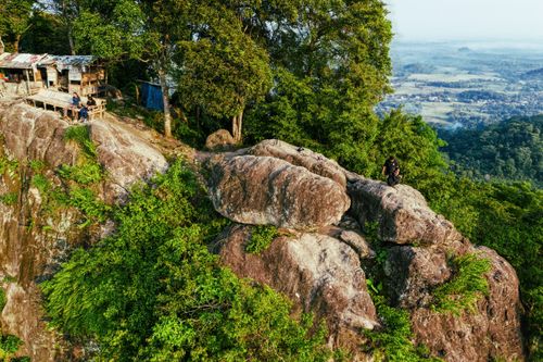 Free Unrecognizable travelers resting on massive rocky cliff near small wooden houses surrounded by lush green trees on sunny day in highland Stock Photo