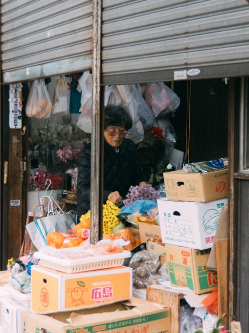 Asian female seller standing at counter of street store with variety of flowers and goods