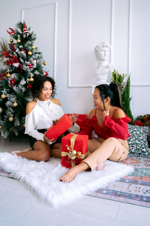 2名妇女坐在绿色圣诞树旁的白色毛皮纺织品上