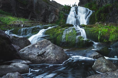 Long exposure of picturesque rapid water cascade streaming through rough rocks with green grass in daylight