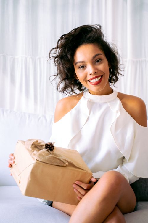 Free Joyful woman sitting indoors holding a wrapped gift box, smiling warmly at the camera. Stock Photo