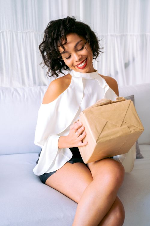 Free Smiling woman in stylish blouse opening a gift box on a sofa, filled with joy and excitement. Stock Photo