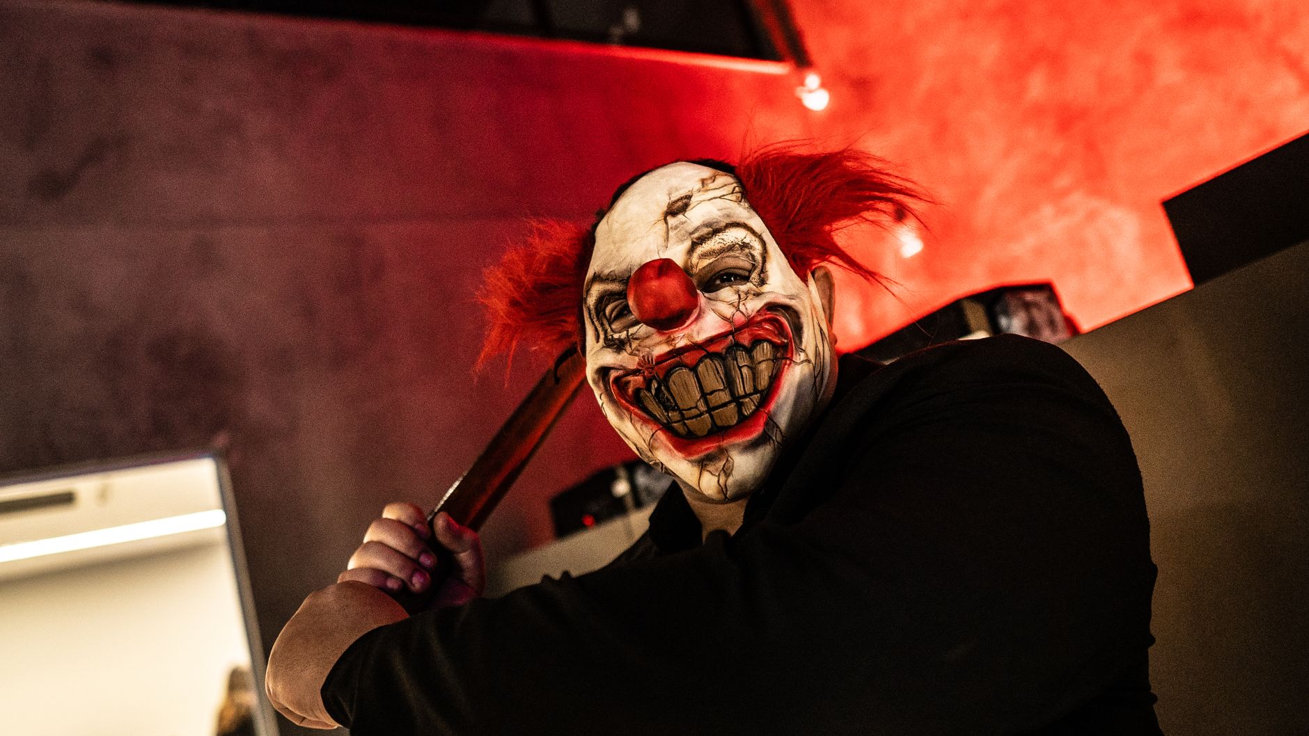 Creepy clown with a frightening mask holding a knife indoors under red lighting.