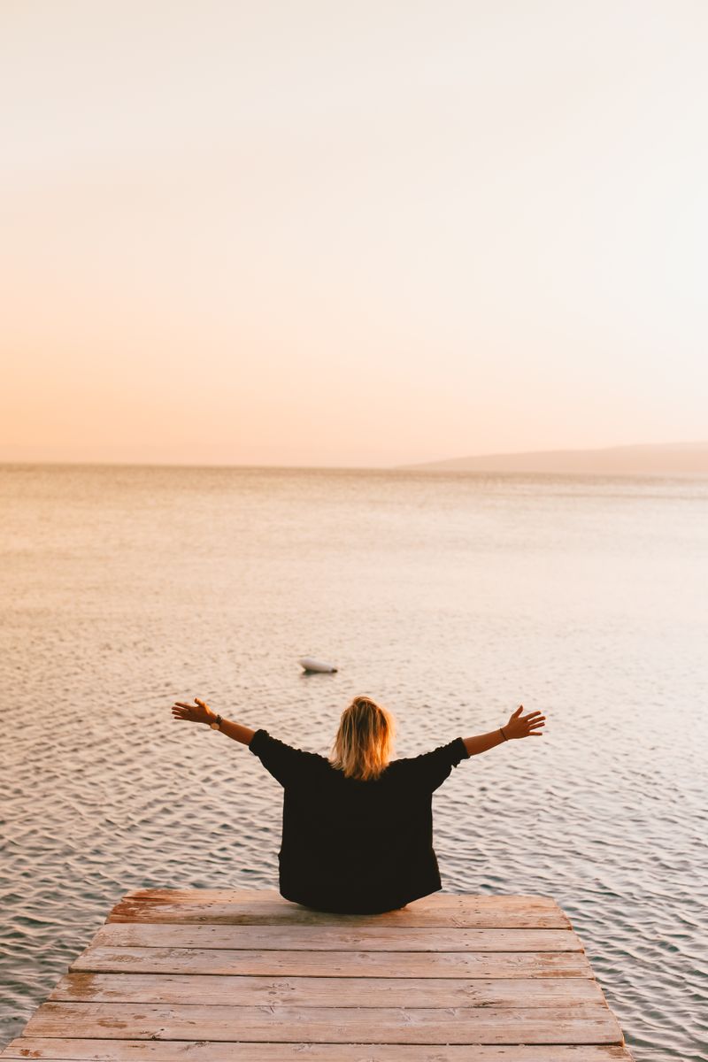 A person sits on a dock with open arms, enjoying a peaceful sunset by the sea.