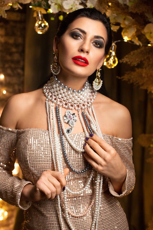 Trendy female with makeup wearing festive accessories and stylish dress with bare shoulders looking at camera while standing room with glittering golden ornaments