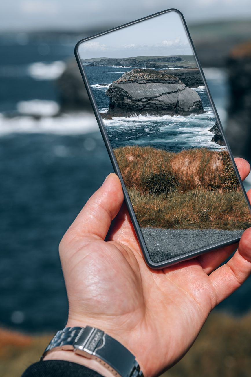 Hand holds smartphone showing rocky ocean cliff with waves crashing below.