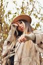 From below of trendy female wearing coat and headwear looking at camera while standing with hand near face with green branches on blurred background