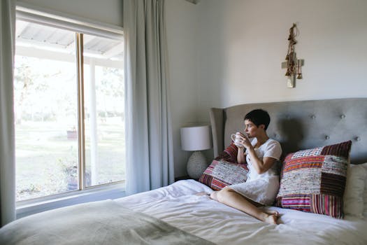 A woman sits in a cozy bedroom with a cup of coffee, gazing through the window.