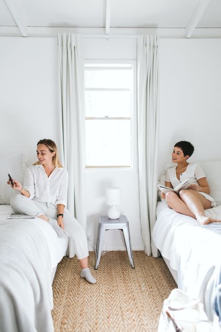Two women relaxing in a bright, minimalist bedroom with white bedding.