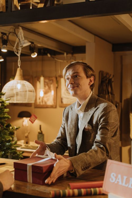 Free Smiling businessman in suit presenting a wrapped gift in a warmly lit shop with Christmas decor. Stock Photo