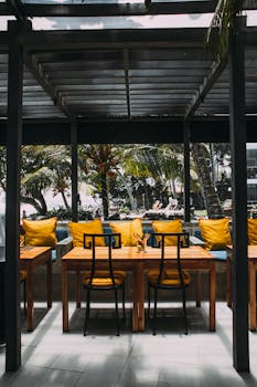 A stylish outdoor setting features wooden tables, yellow pillows, and tropical greenery, perfect for a relaxed dining experience.