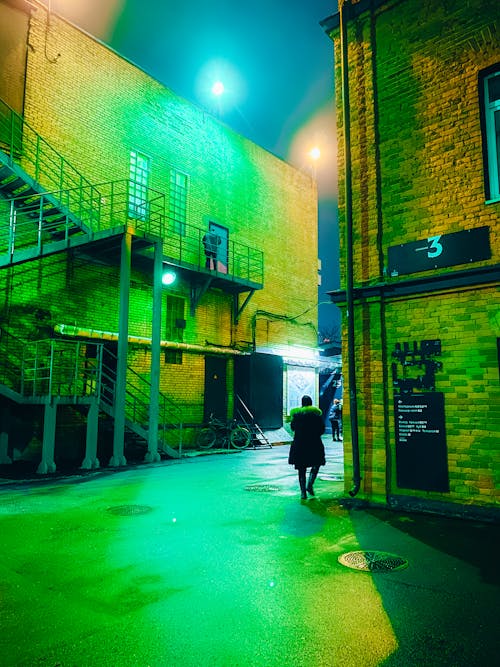 Unrecognizable person walking between industrial buildings with staircase and signboards and green neon light in megapolis at night