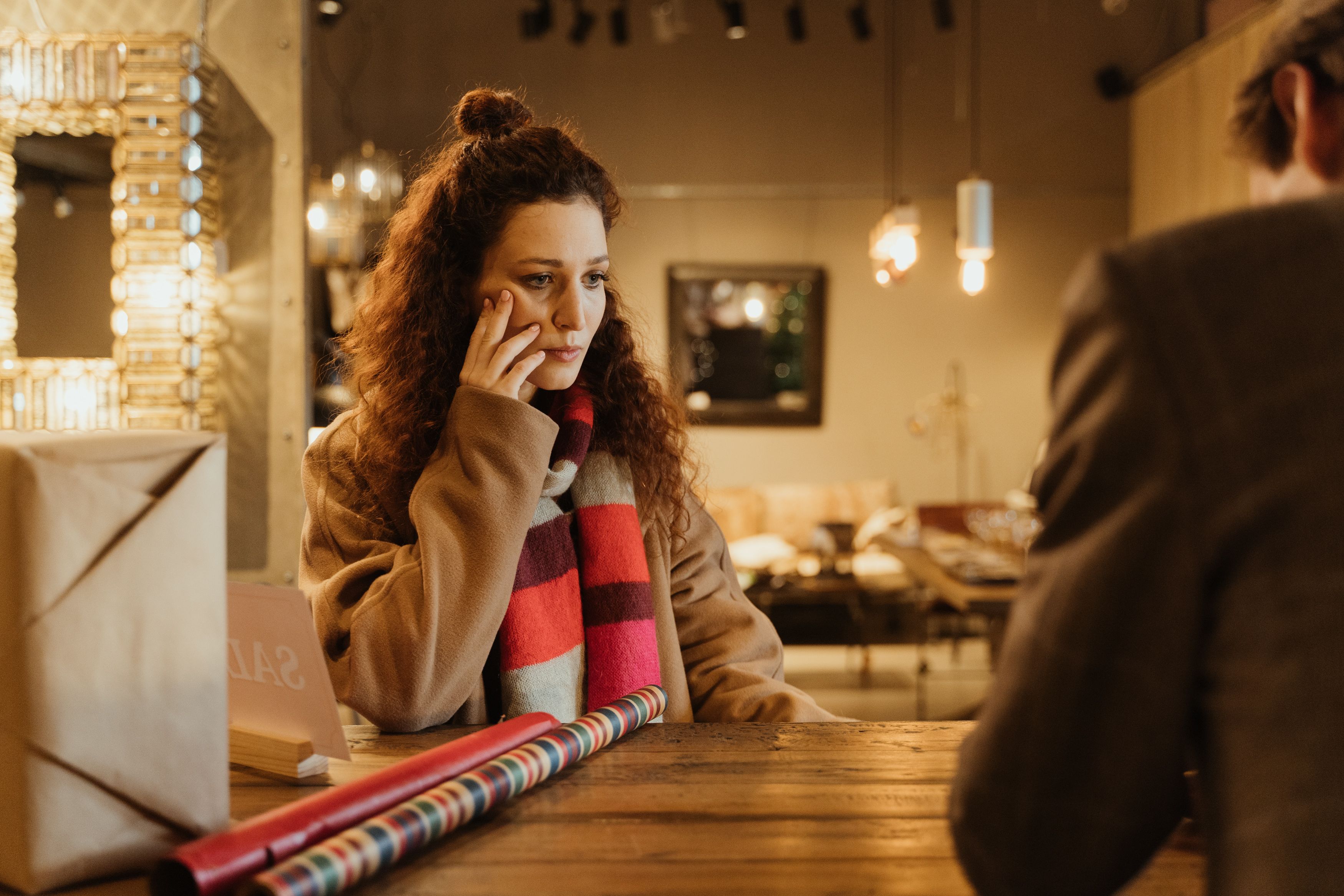 A woman in a cozy cafe, sitting thoughtfully at a table with gift wrapping materials.
