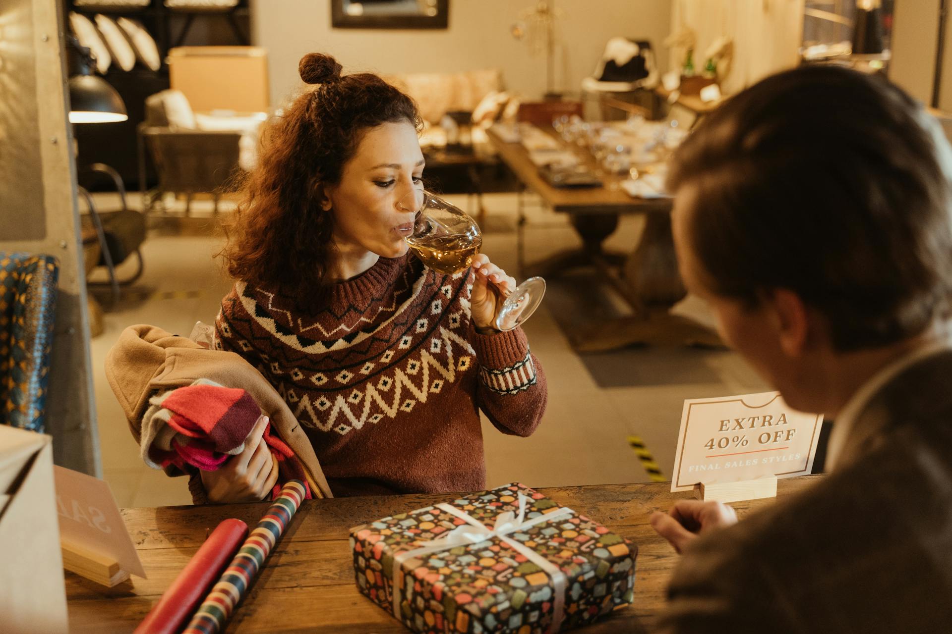 A woman enjoying wine at a shop while selecting holiday gifts with a 40% discount offer visible.