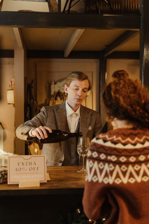 Free A man in a suit pours wine for a customer inside a cozy store with a discount sign visible. Stock Photo