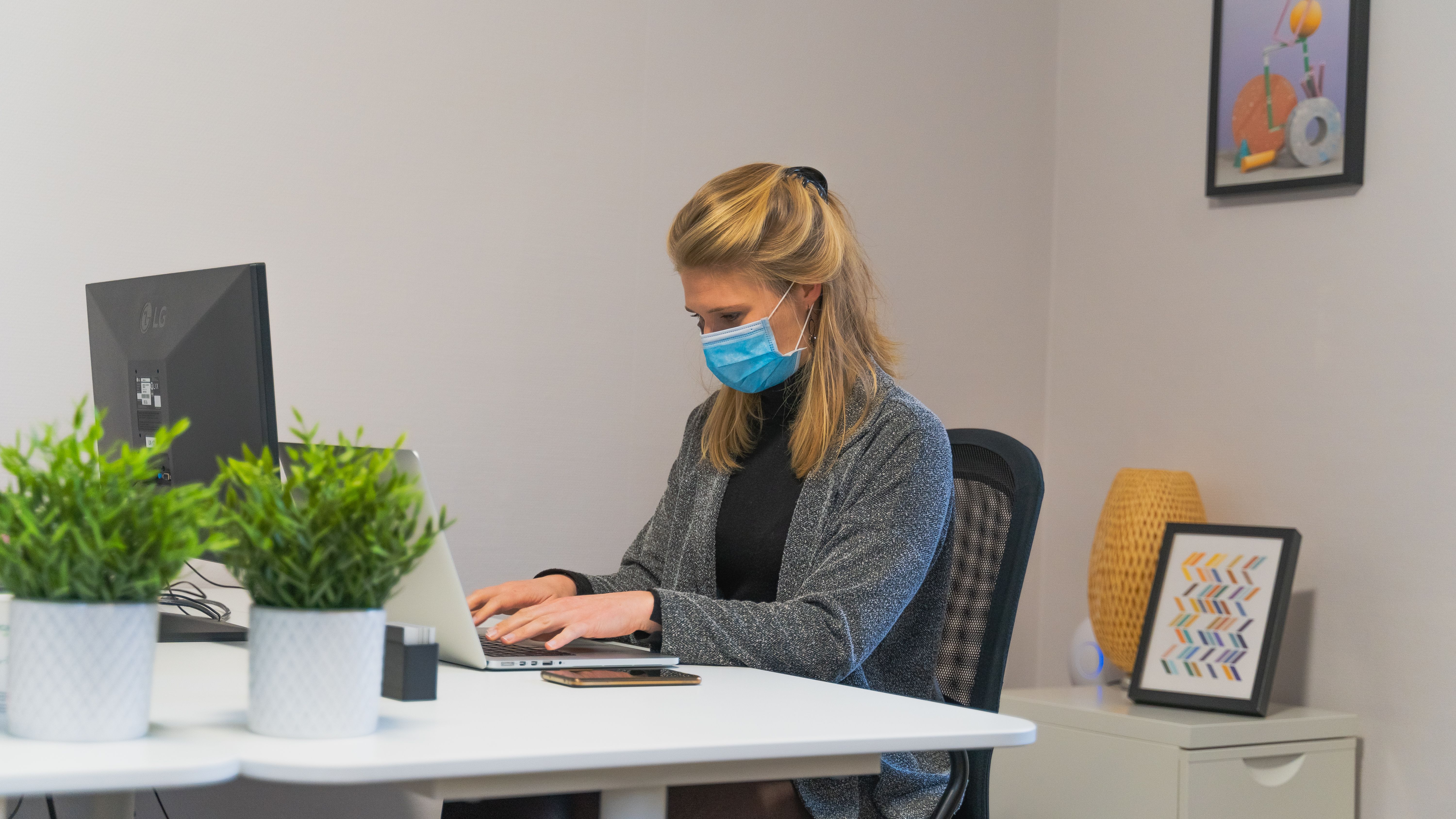 A woman in a face mask working on her laptop at an indoor office desk.