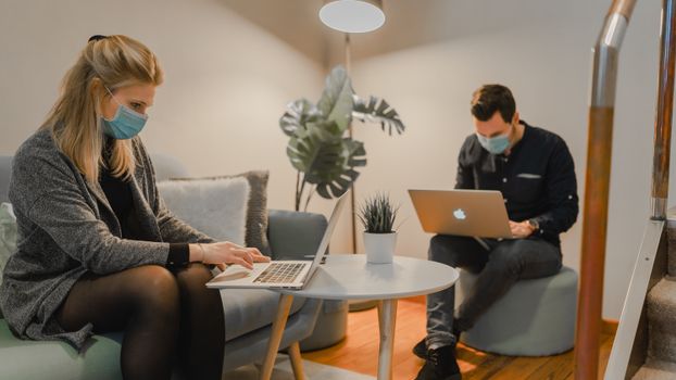 Two people wearing masks working on laptops in a cozy office space with home plants.