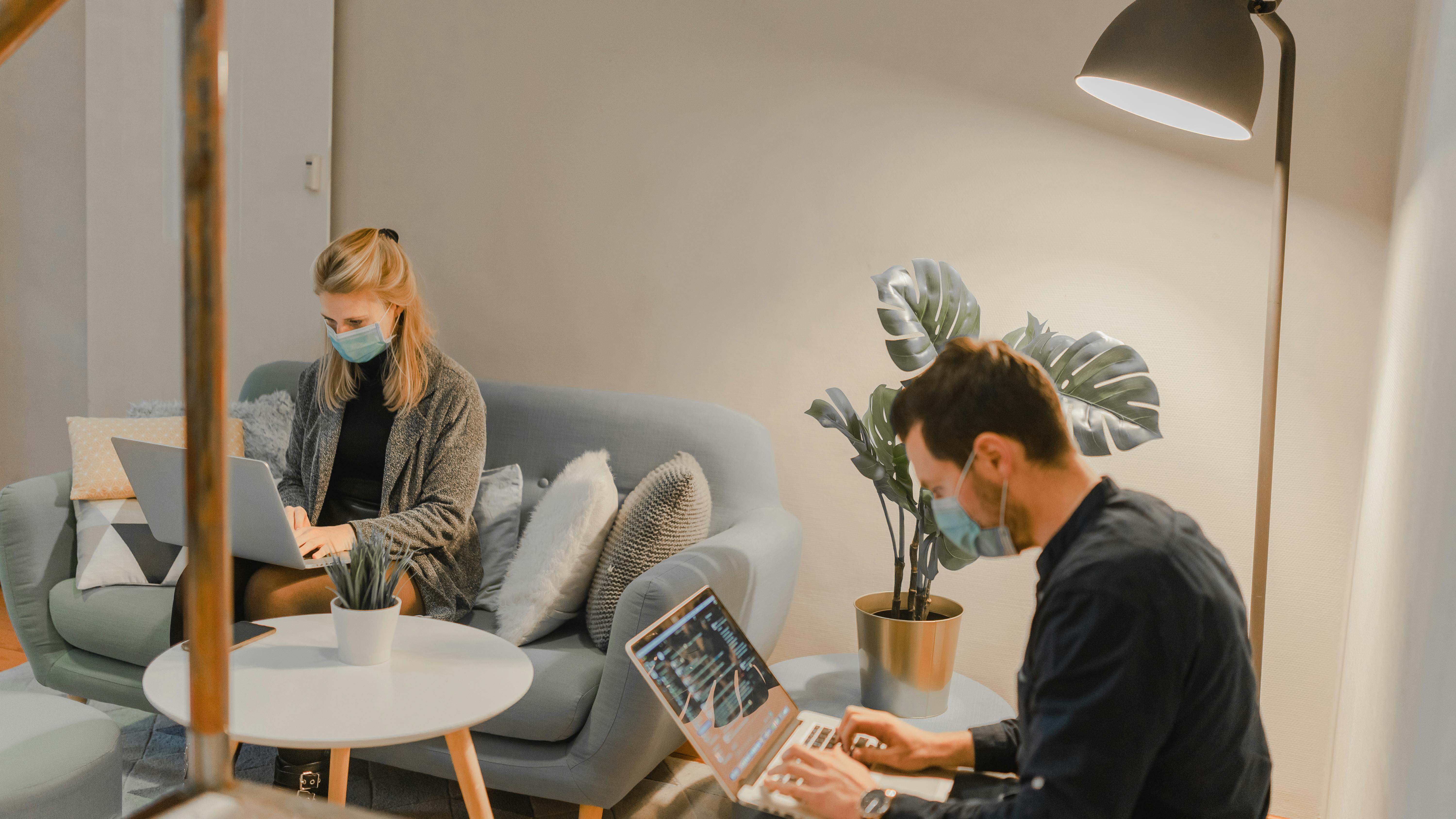 Two employees with face masks working on laptops in an office lounge.