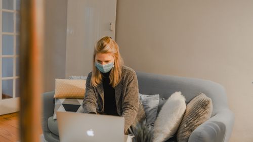 Free Woman working remotely from home on a laptop, wearing a face mask on a cozy couch during pandemic. Stock Photo