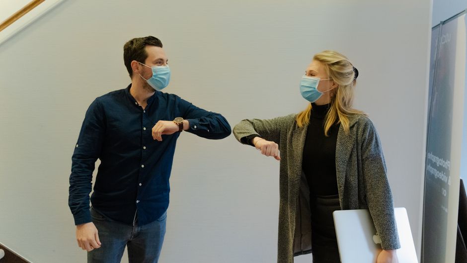 Dos compañeros se saludan con un golpe de codo usando mascarillas en un entorno de oficina.