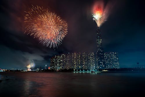 Free Night dark cloudy sky with colorful flashing fireworks above river and modern town with skyscrapers Stock Photo