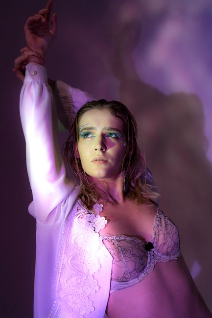 Dreamy young lady in white shirt and bra with eyeshadows looking away while standing in studio on purple background with raised arms