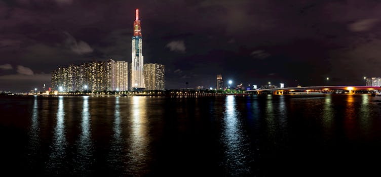 Breathtaking view of dark water near Ho Chi Minh City in Vietnam with glowing skyscrapers and Landmark 81 building near bridge at night under black cloudy sky