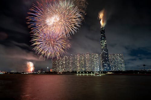 Illuminated skyscrapers with tall building named Landmark 81 in Vietnam in Ho Chi Minh City near black river with colorful fireworks under dark cloudy sky at night