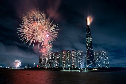 Illuminated skyscrapers in Vietnam in Ho Chi Minh City with tall building named Landmark 81 near black water and colorful fireworks in twilight under dark cloudy sky