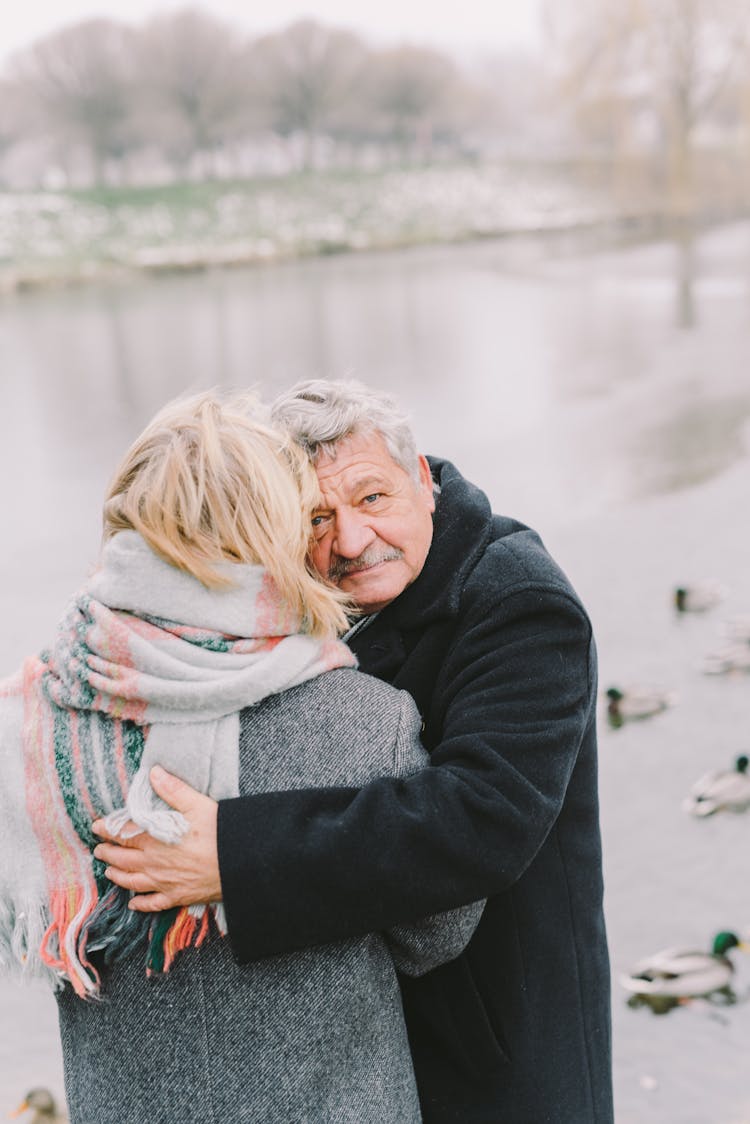 Elderly Man In Black Coat Hugging A Woman Near Lake