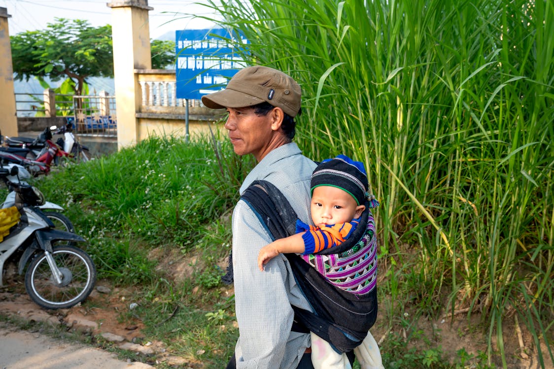 Free Back view content Asian male in casual wear and cap carrying cute baby on back carrier while standing in lush village Stock Photo