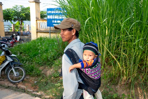 Free Back view content Asian male in casual wear and cap carrying cute baby on back carrier while standing in lush village Stock Photo