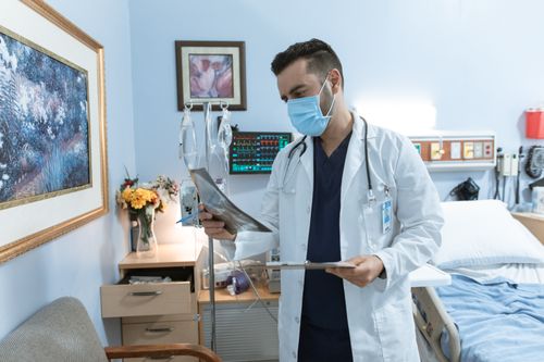 Free A male doctor wearing a face mask reviews an X-ray in a hospital room, focusing on patient diagnosis. Stock Photo