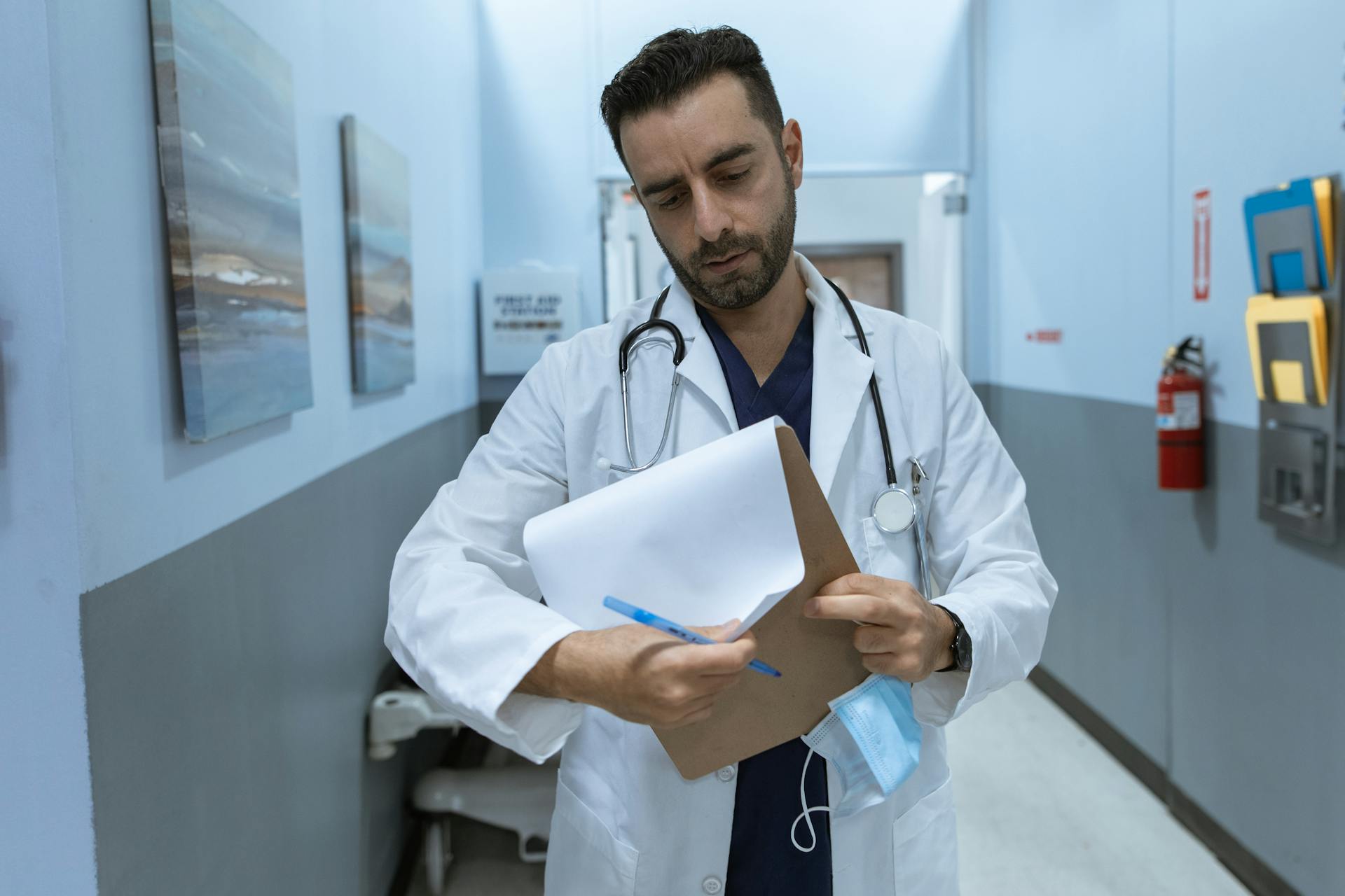 A doctor in a lab coat reviews a medical chart in a hospital hallway.