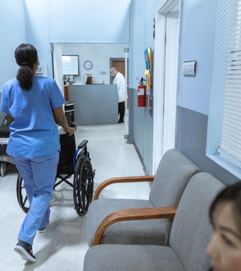 A nurse in scrubs pushes a wheelchair through a well-lit hospital corridor, attending to patient care.