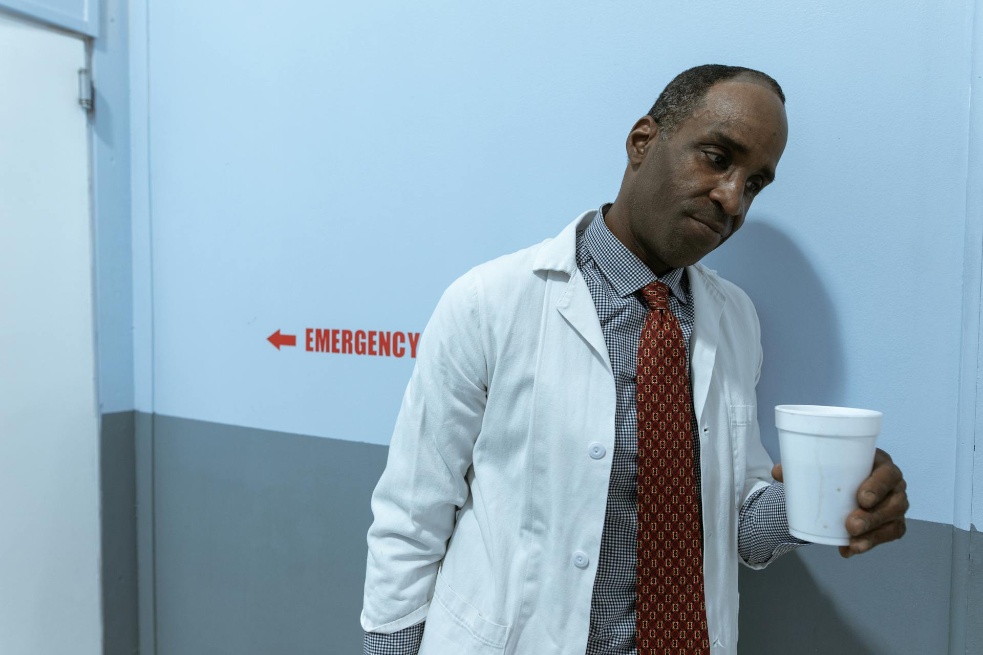 A healthcare worker in a lab coat holding a cup in a hospital corridor by the emergency section.