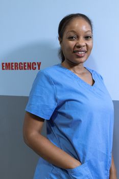 A cheerful nurse standing in front of an emergency room wall, wearing blue scrubs.