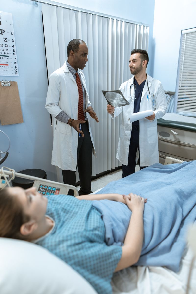 A Black physician reviews an X-ray with a colleague at a patient's hospital bedside. Hospital stays and surgery are among the most common settings for a blood clot to form.