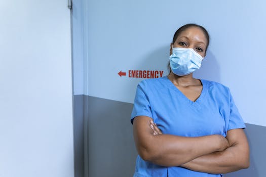Nurse in blue scrubs and face mask in hospital corridor with emergency sign.