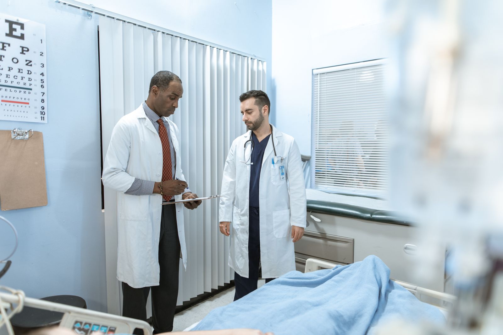 A Black physician reviews a patient's chart with a colleague in a hospital exam room, the kind of urgent evaluation a TIA demands within 48 hours.