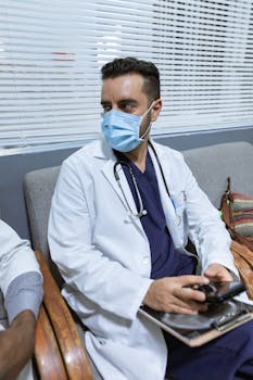 Doctor in lab coat and face mask, sitting with smartphone and clipboard, ready for consultation.