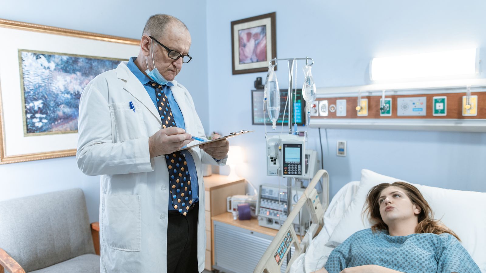 Doctor in white coat checking patient chart in hospital room with medical equipment.