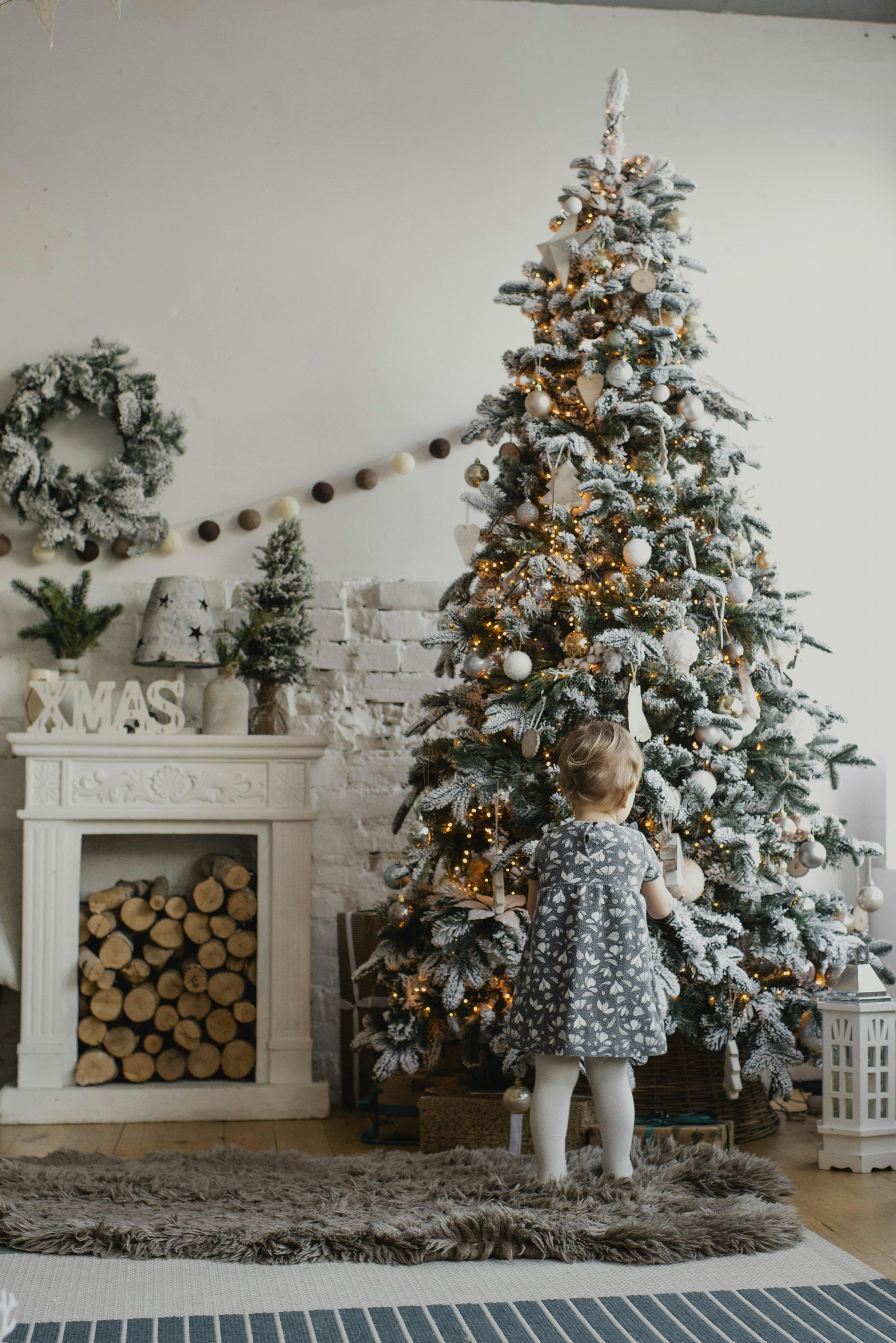 A young child in a festive dress admires a beautifully decorated Christmas tree indoors, capturing the holiday spirit.